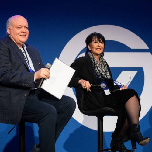 Two people are seated on stage in front of a blue backdrop with a GVSU logo. The person on the left, wearing a dark blazer and holding a microphone and papers. Person on the right, wearing a black outfit, scarf, and heeled boots.